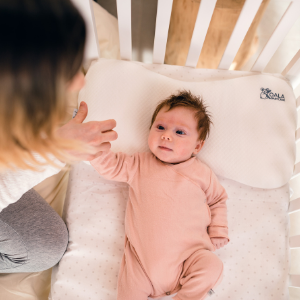 Maman tient la main de sa fille couchée dans berceau avec oreiller bébé blanc Plagiocéphalie Maxi